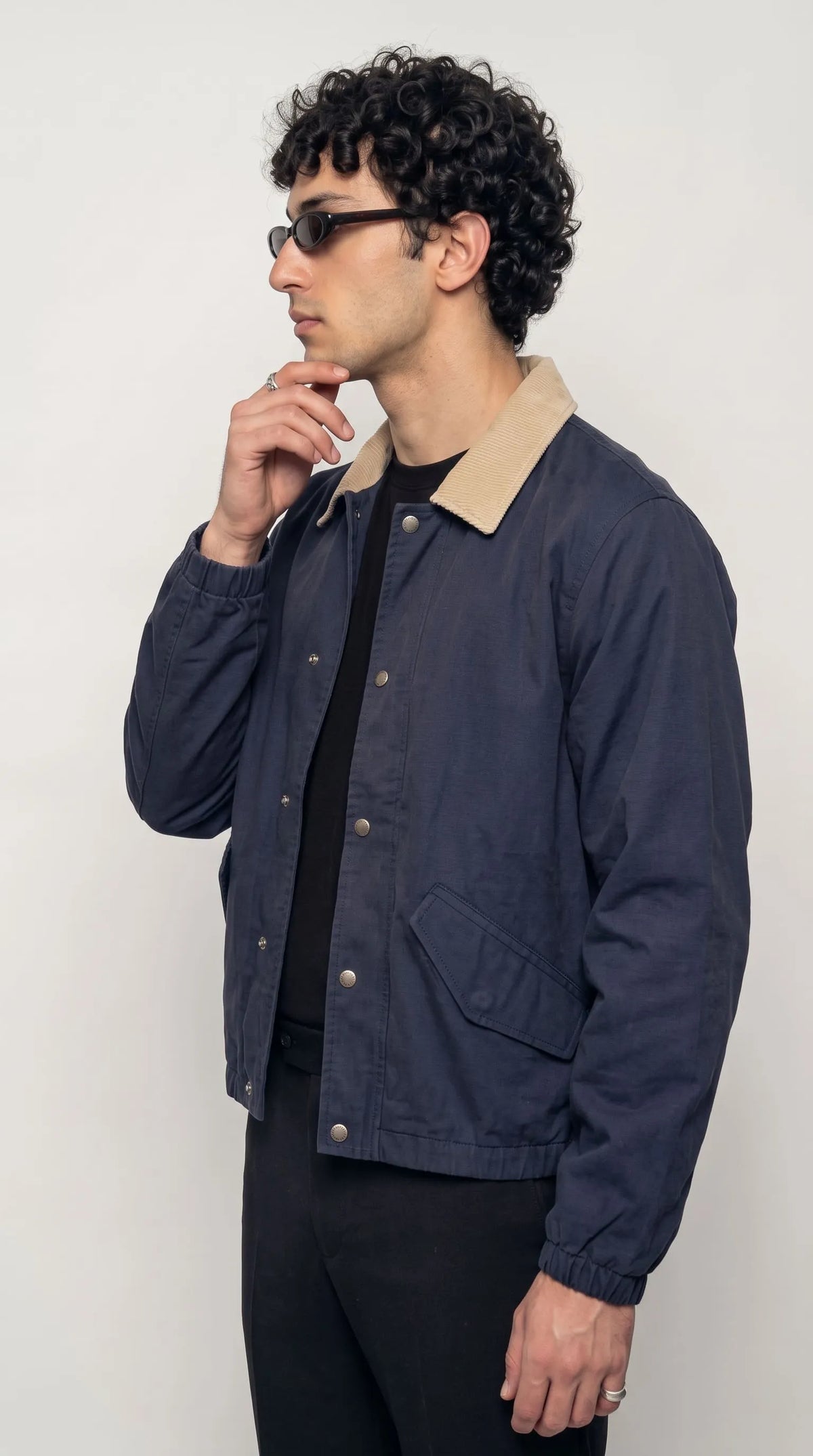 Dark navy cotton canvas blouson jacket with a light beige contrast collar, full-length silver snap-button fastening, elasticated sleeve cuffs and practical slash pockets, paired with a black crew-neck T-shirt for understated contemporary urban fashion.