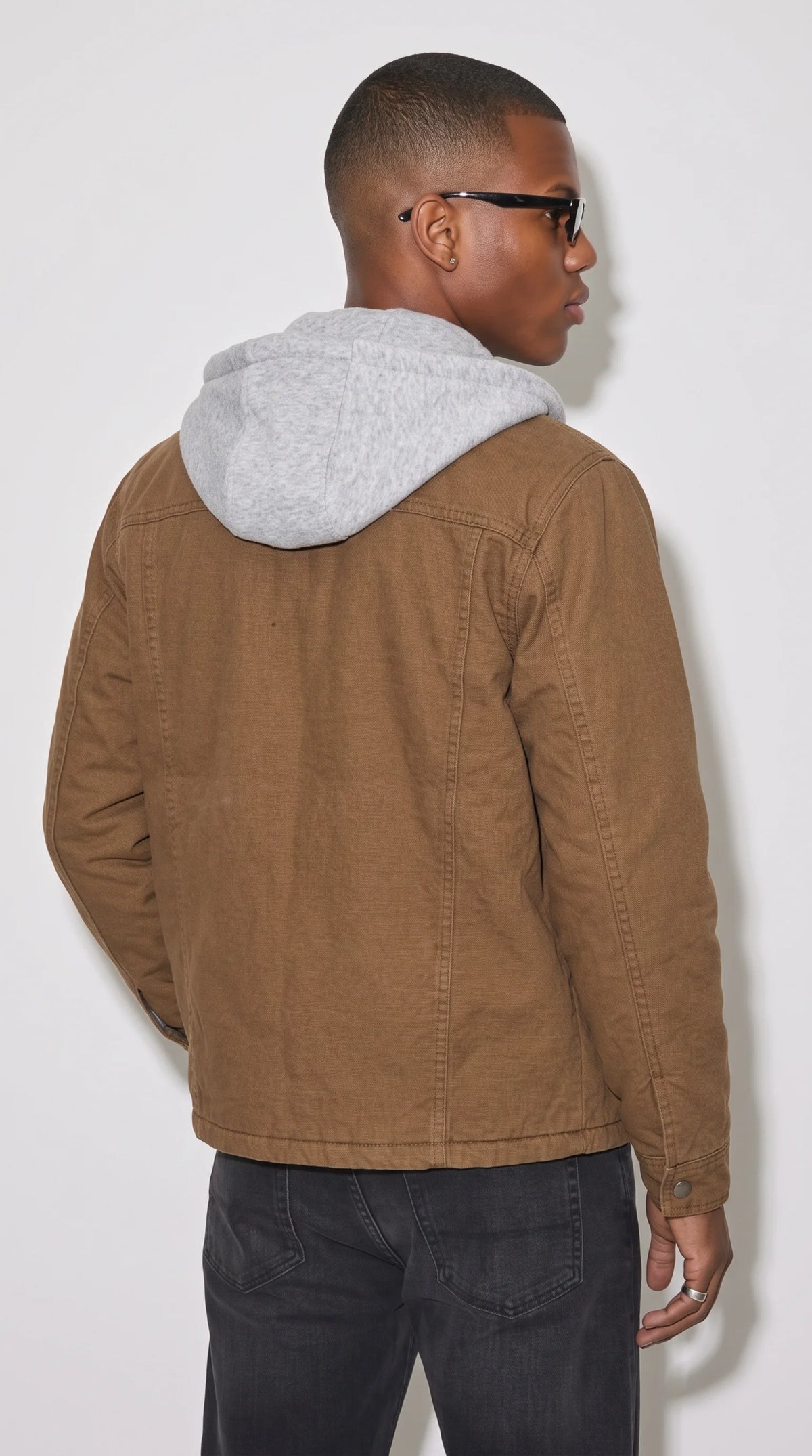Dark tan heavyweight cotton work jacket with a contrasting grey jersey hood, horizontal shoulder yoke, vertical back-panel seams and a straight hem, showcasing its sturdy construction and relaxed silhouette for heritage-inspired workwear and everyday outdoor style.