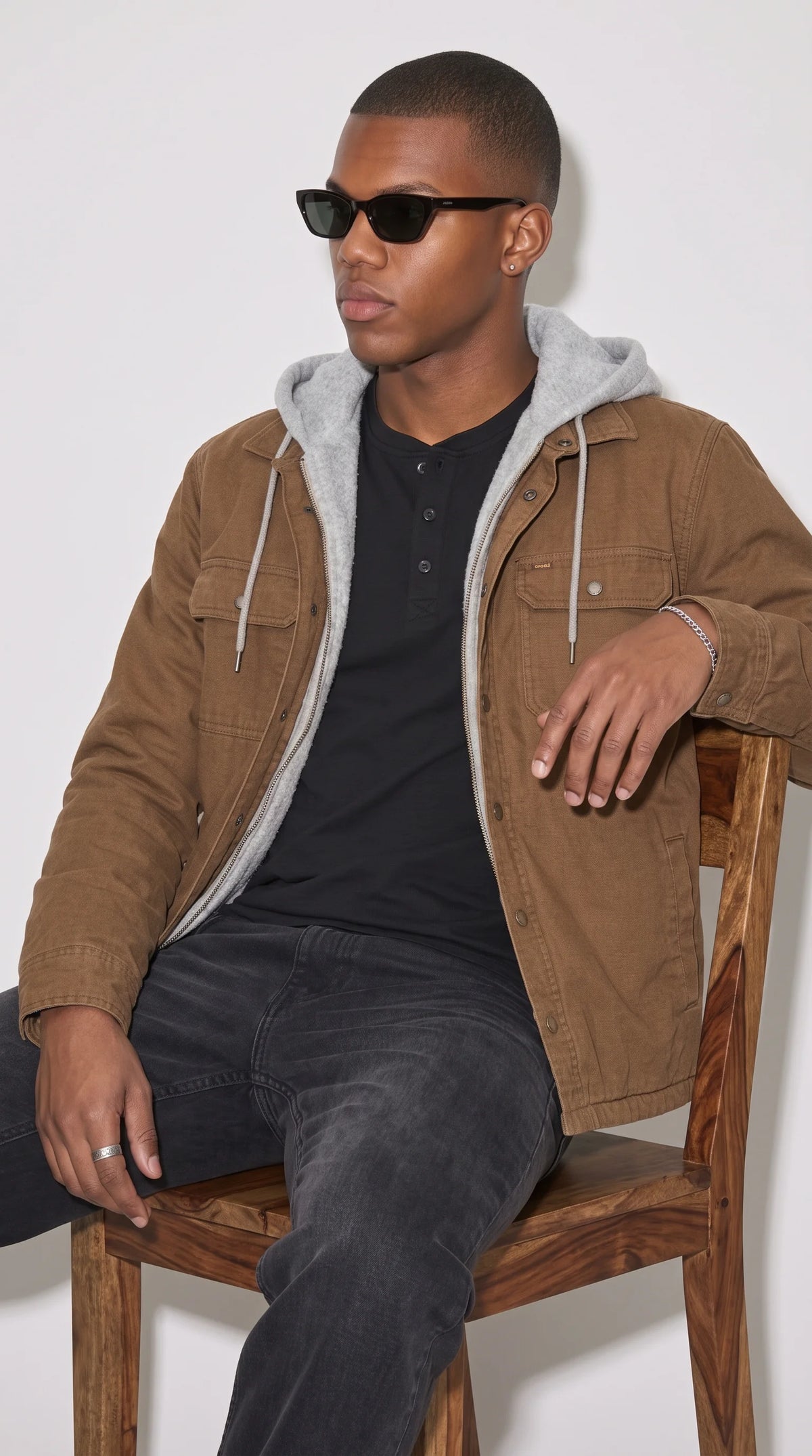Dark tan cotton workwear jacket styled with a black henley shirt and dark denim, showcasing its contrasting grey hood, twin flap chest pockets, textured fabric and relaxed shoulder fit for an understated monochrome outfit with rugged street-style character.