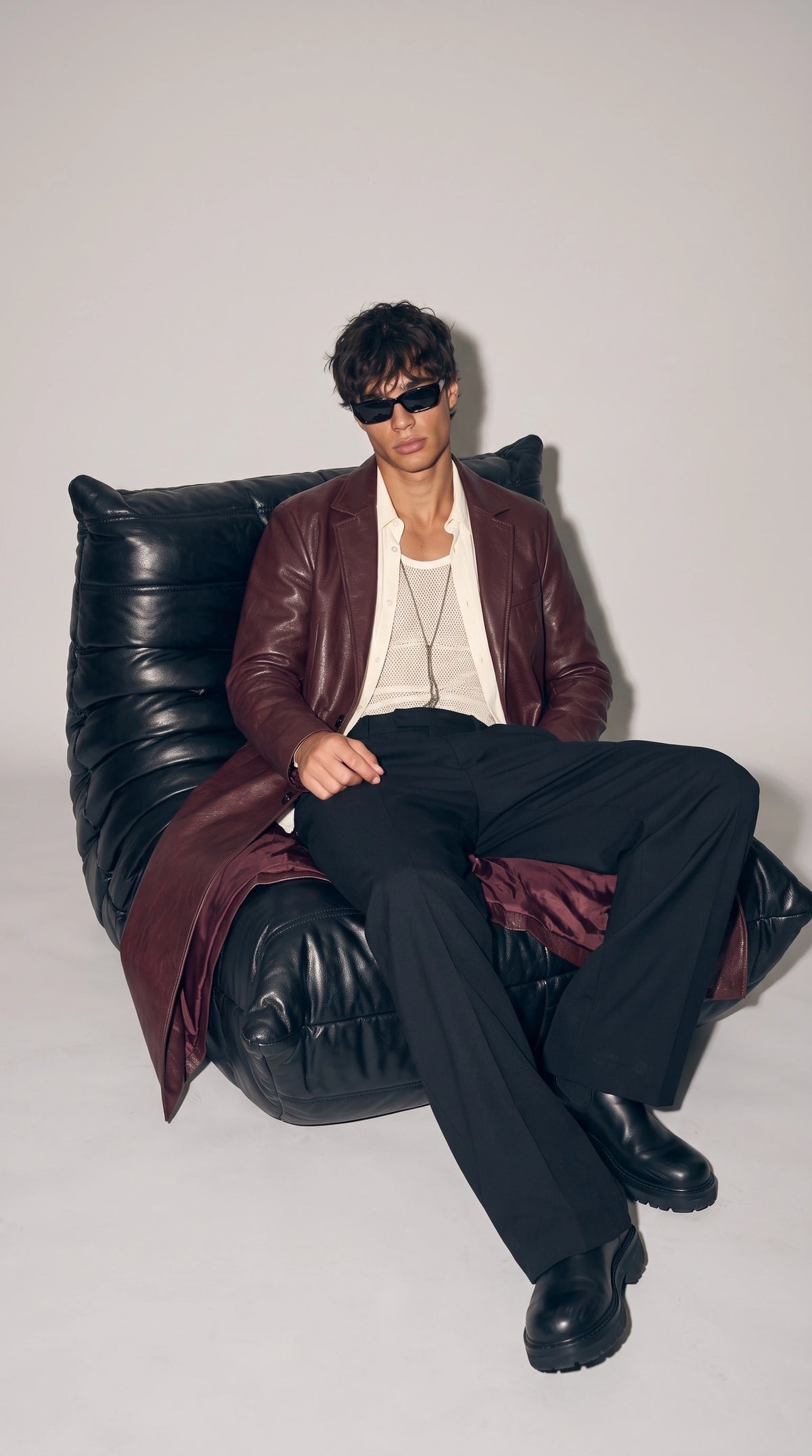 Men’s dark burgundy leather long coat worn open over an ivory button-up shirt and textured mesh top, paired with relaxed black trousers and chunky black footwear while seated on a black leather sofa. The flowing coat length and rich wine-coloured leather create a confident retro-luxury mood, ideal for editorial menswear, evening outfits and contemporary statement dressing.