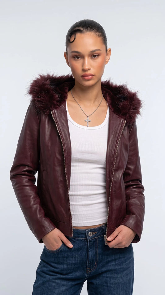Deep wine-red genuine leather hooded jacket with a voluminous dark faux-fur trim, full-length metal zipper and relaxed contemporary silhouette, styled over a white ribbed tank top with dark-blue jeans and a silver cross necklace. The rich burgundy leather and dramatic dark hood create a striking statement outfit that blends modern streetwear with sophisticated cold-weather layering.
