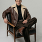 Man wearing a brown leather jacket with fur collar sitting on a chair against a gray background