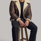 Man wearing a brown leather jacket with white fur lining, sitting on a wooden stool against a plain background.