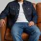 Man wearing a denim jacket and jeans sitting on a brown leather couch against a gray background
