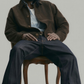 Man sitting on a chair wearing a brown coat and dark pants against a plain background