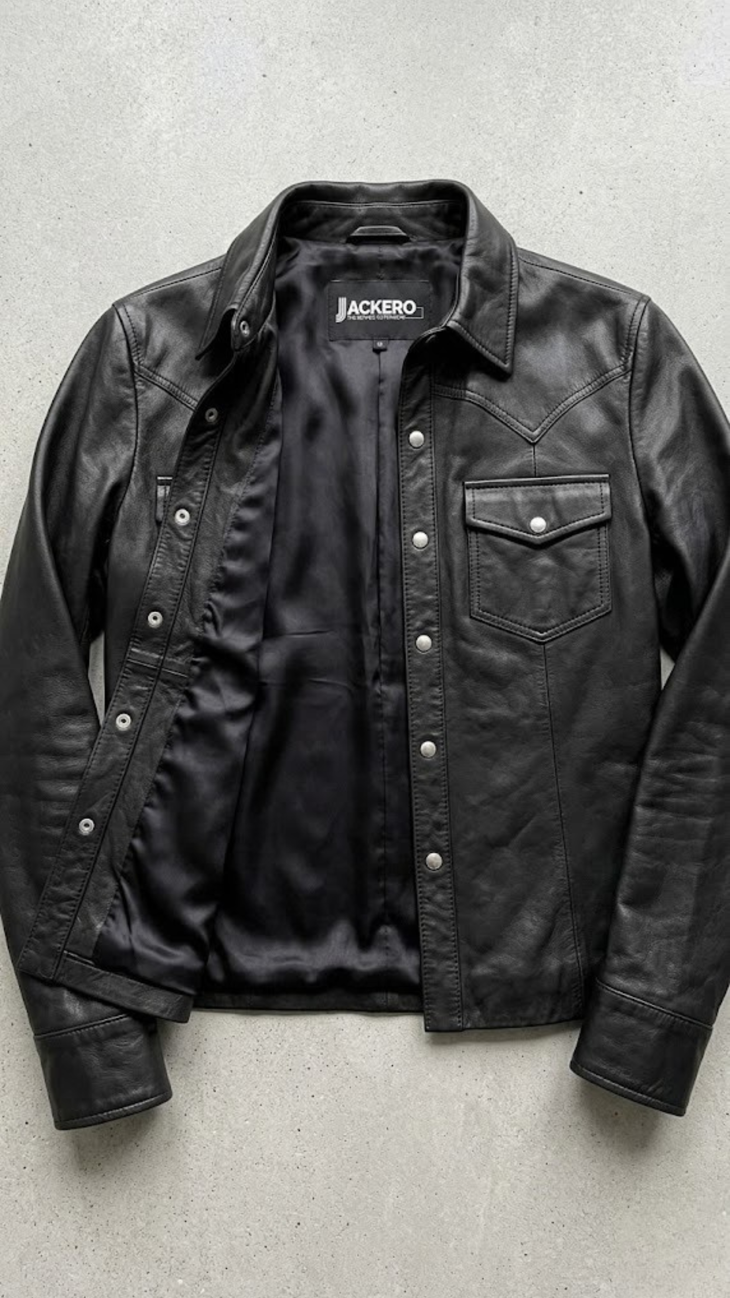Jackero jacket flat lay photography of a high-end black leather shirt-style trucker coat displayed on a solid grey backdrop.