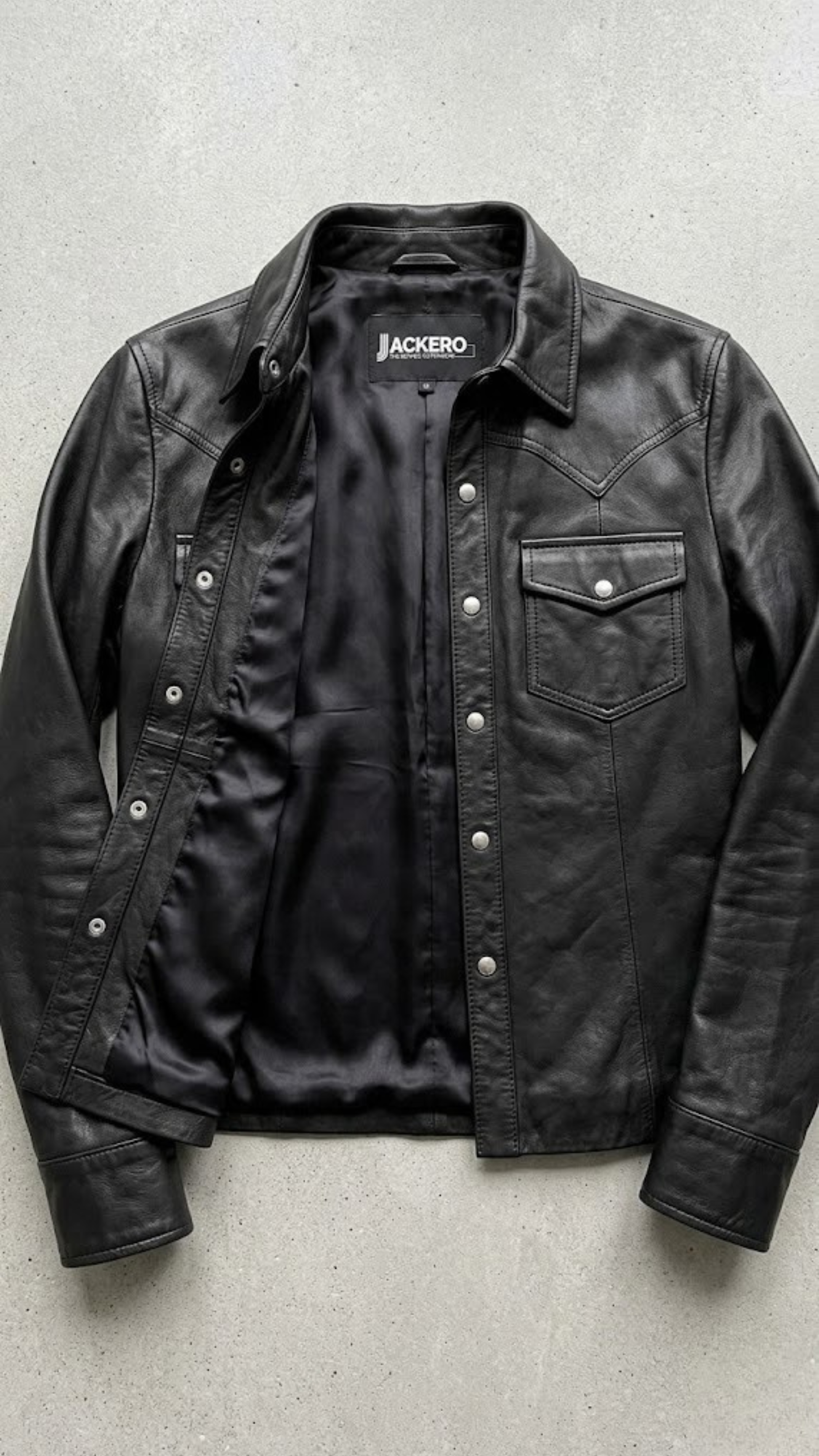 Jackero jacket flat lay photography of a high-end black leather shirt-style trucker coat displayed on a solid grey backdrop.