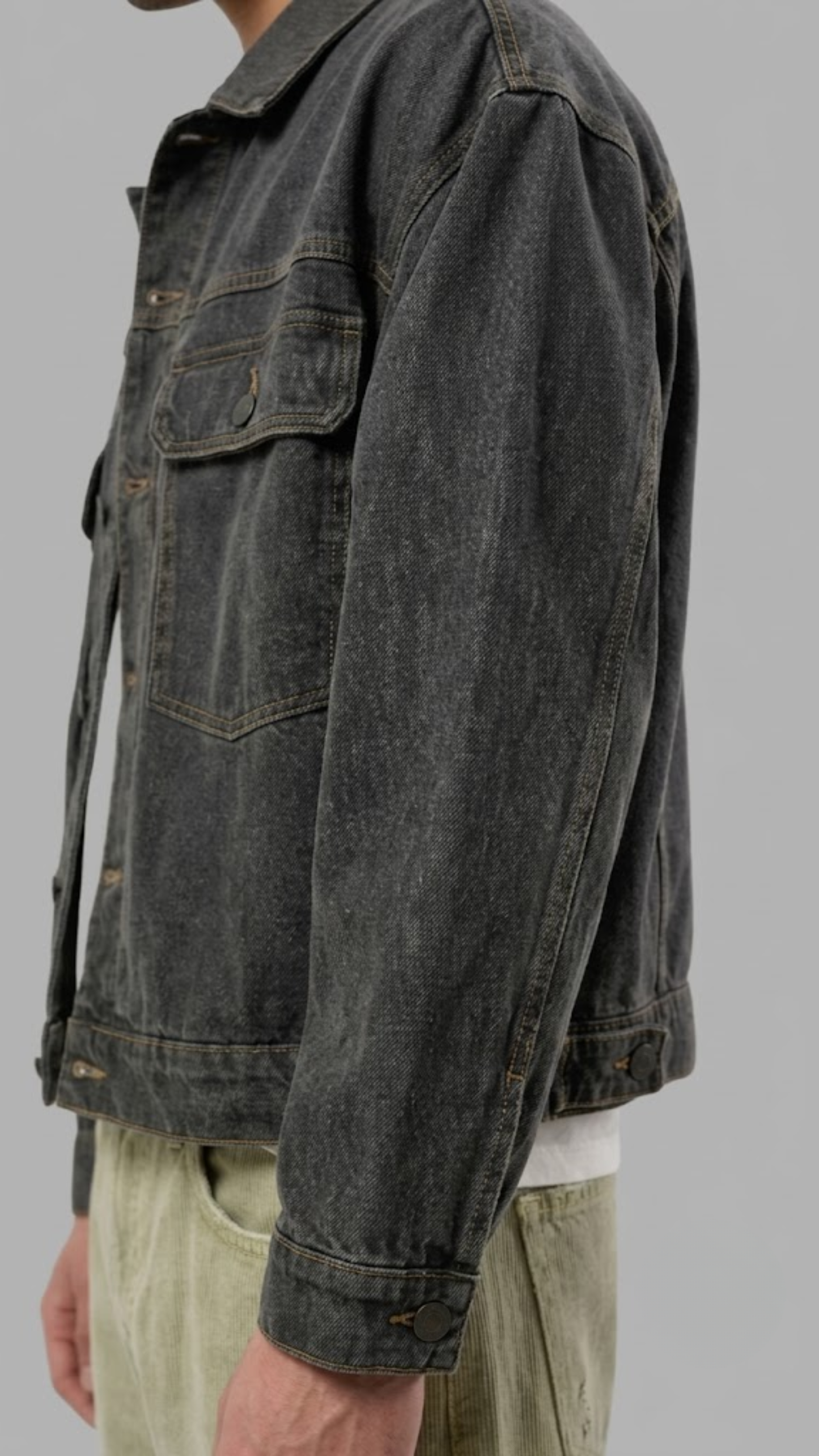 A close-up of a washed dark charcoal denim jacket along the side, shown against a solid gray background.