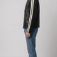 A model with curly blonde hair stands in profile wearing a black with white strips leather jacket, gray washed jeans, and brown sneakers against a neutral gray studio background.