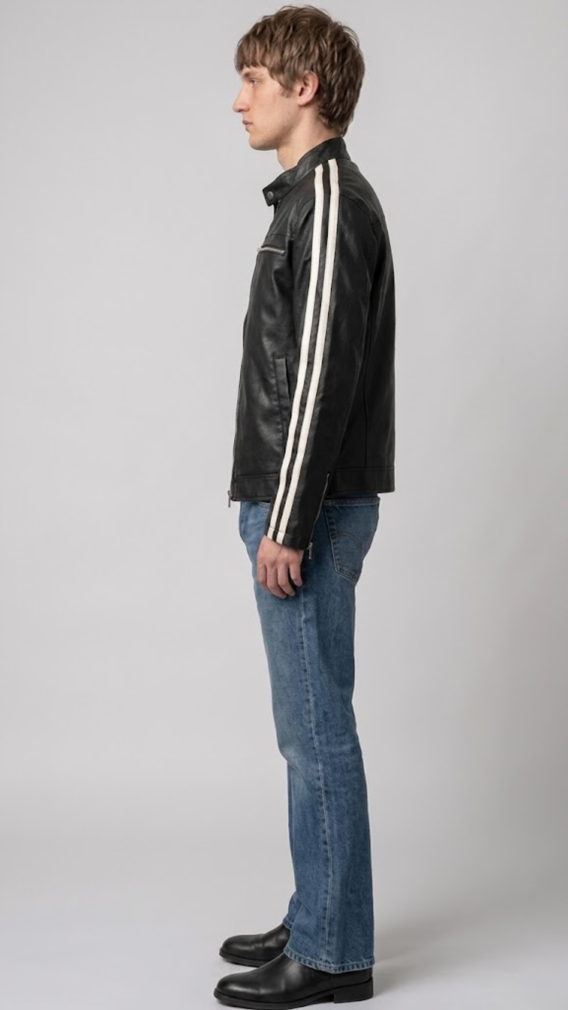 A model with curly blonde hair stands in profile wearing a black with white strips leather jacket, gray washed jeans, and brown sneakers against a neutral gray studio background.