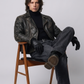 Man sitting on a wooden stool wearing a leather jacket and black gloves against a white background