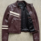 A dark burgundy leather jacket with white strips, laid flat on a textured gray concrete background. The jacket features multiple front flap pockets and a zippered chest pocket, with the JACKEDO brand name visible on the inner neck label.