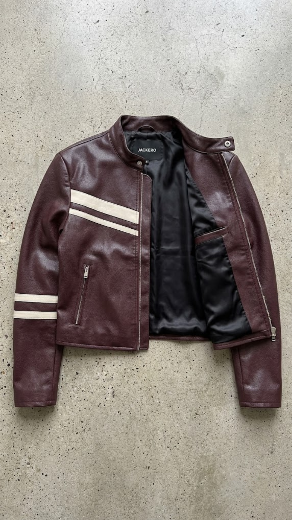 A dark burgundy leather jacket with white strips, laid flat on a textured gray concrete background. The jacket features multiple front flap pockets and a zippered chest pocket, with the JACKEDO brand name visible on the inner neck label.