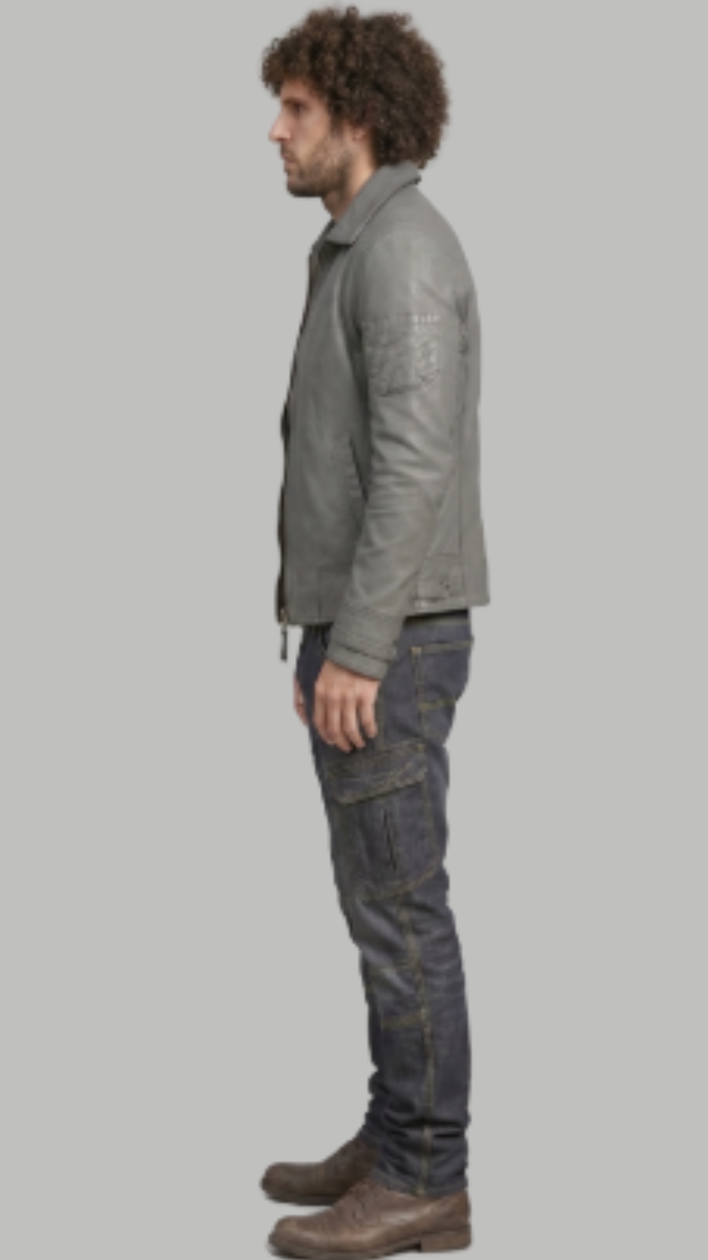 A man shown in profile wearing a gray leather jacket, dark gray cargo jeans, and brown boots, standing against a solid gray background.