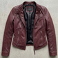 A burgundy leather moto jacket with a black interior lining, laid flat on a textured gray concrete surface. The brand name 'JACKIE' is visible on the inner neck label.