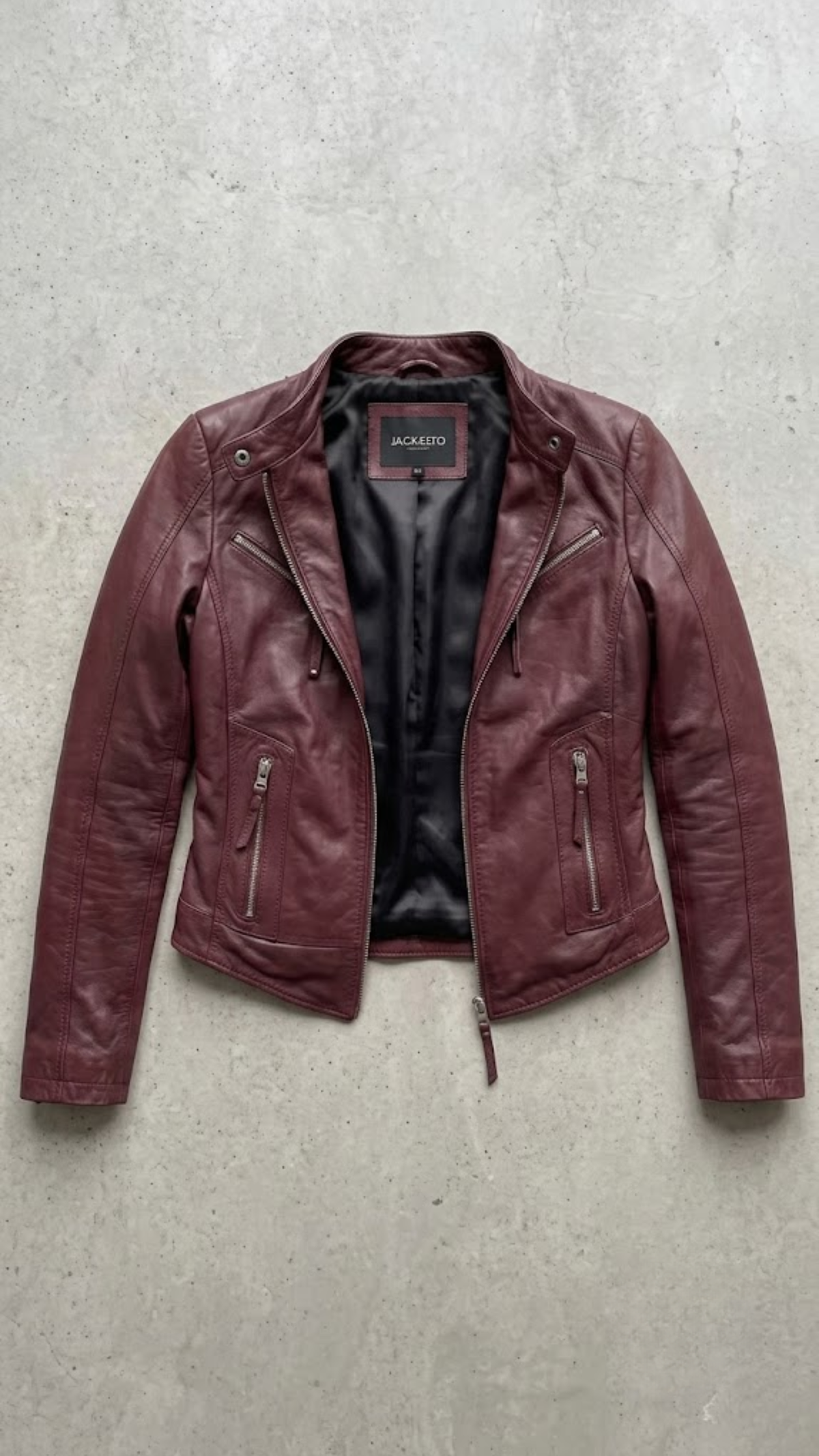 A burgundy leather moto jacket with a black interior lining, laid flat on a textured gray concrete surface. The brand name 'JACKIE' is visible on the inner neck label.