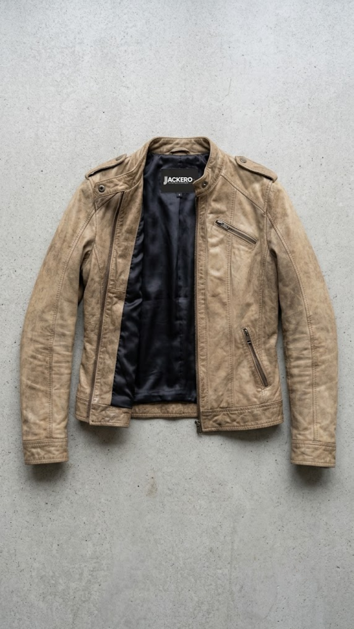 A cropped brown faux leather jacket with cream-colored diagonal stripes on the chest and sleeves, laid flat on a textured gray concrete surface. The brand name 'JACKERO' is visible on the inner neck label.