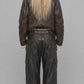 A model shown from the back wearing a dark brown leather jacket with a shearling-style collar, dark wide-leg cargo pants with chain detailing at the back, and a black patterned bandana with long blonde hair tied underneath, set against a plain gray studio background.