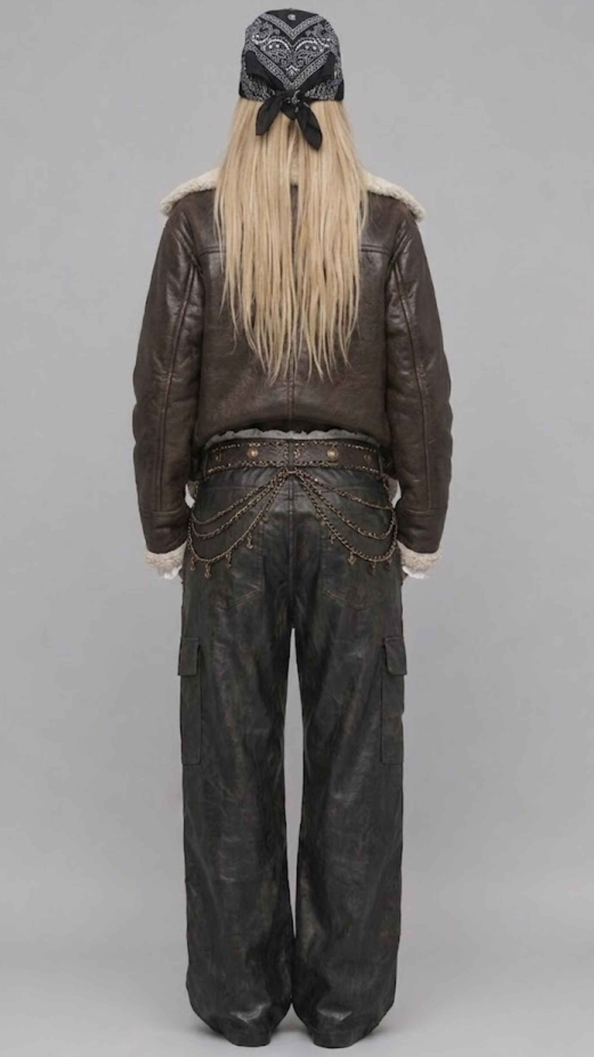 A model shown from the back wearing a dark brown leather jacket with a shearling-style collar, dark wide-leg cargo pants with chain detailing at the back, and a black patterned bandana with long blonde hair tied underneath, set against a plain gray studio background.