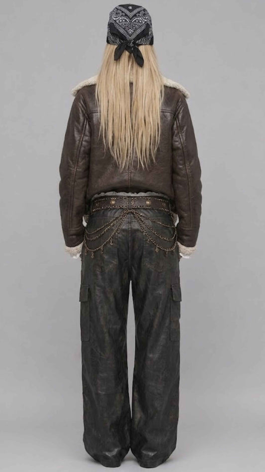 A model shown from the back wearing a dark brown leather jacket with a shearling-style collar, dark wide-leg cargo pants with chain detailing at the back, and a black patterned bandana with long blonde hair tied underneath, set against a plain gray studio background.
