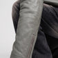 Close-up of a person wearing a light gray leather jacket with a diagonal zip pocket on the forearm, paired with dark gray jeans and black boots.