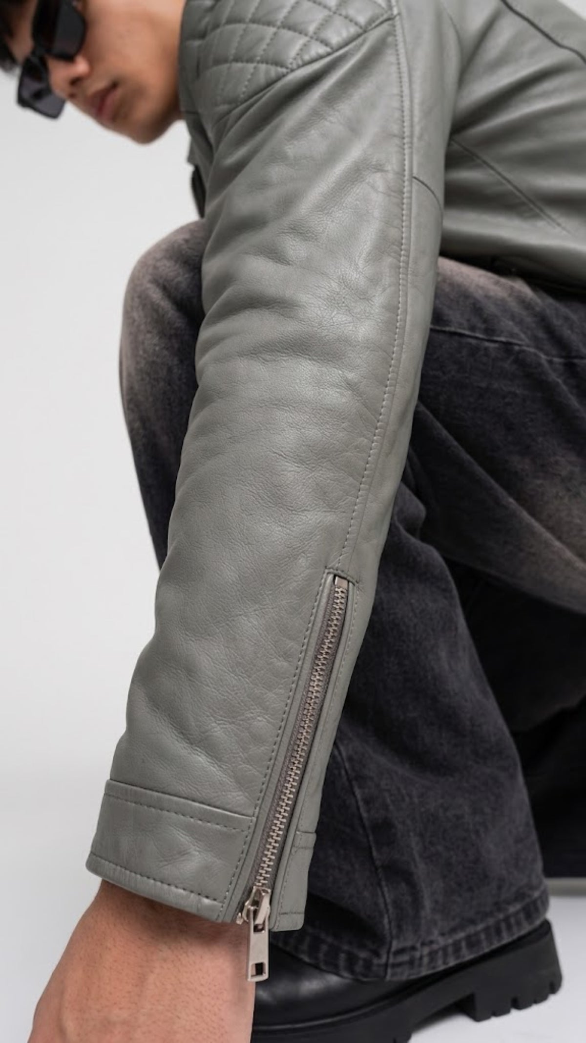Close-up of a person wearing a light gray leather jacket with a diagonal zip pocket on the forearm, paired with dark gray jeans and black boots.