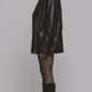 A woman shown in profile wearing a black leather jacket, sheer black tights, and black knee-high boots against a plain light gray studio background.