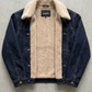 A dark blue denim jacket with a thick beige sherpa fleece lining, laid flat on a textured gray concrete surface. The brand name 'JACKERO' is visible on the inner neck label.