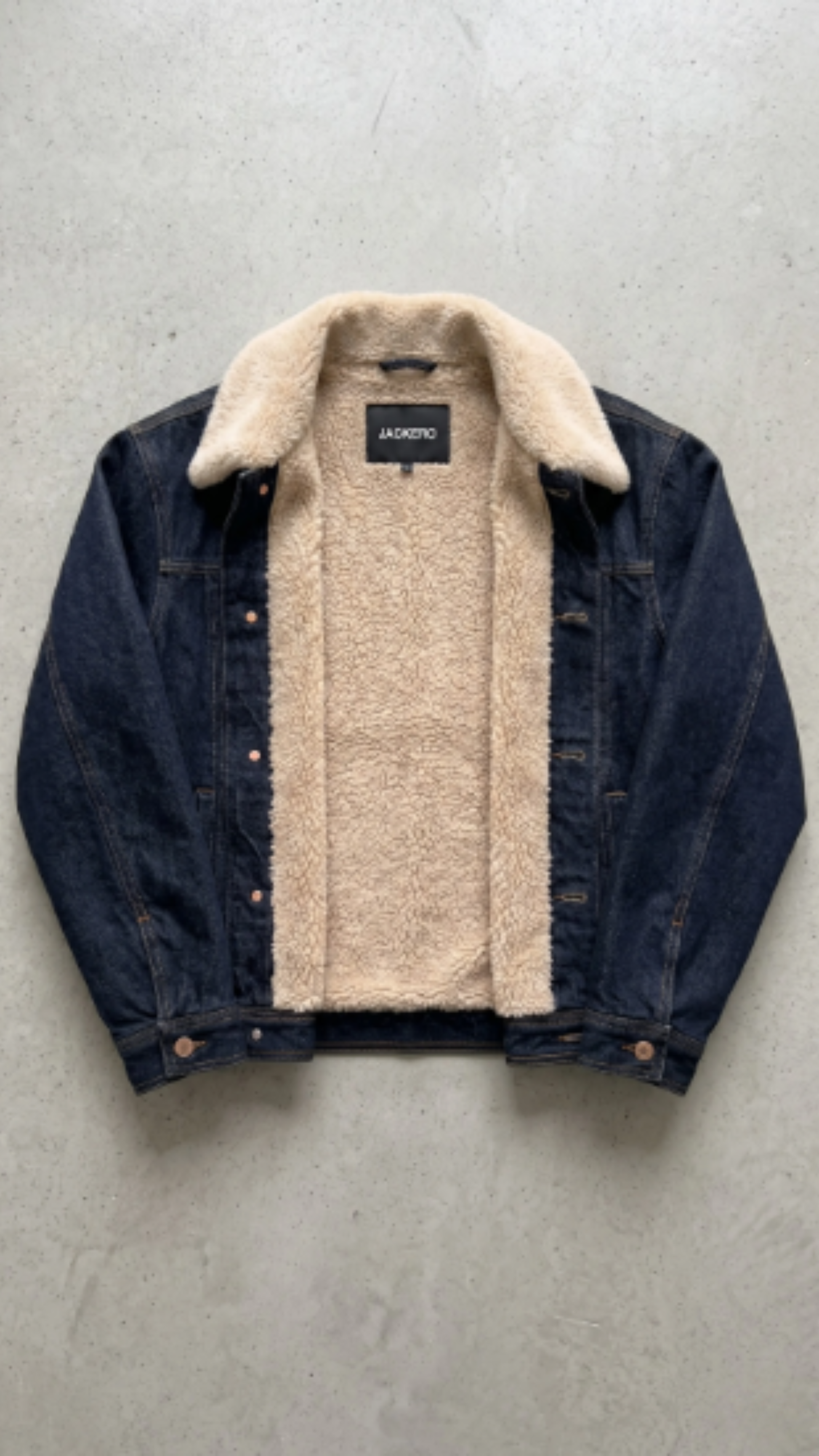 A dark blue denim jacket with a thick beige sherpa fleece lining, laid flat on a textured gray concrete surface. The brand name 'JACKERO' is visible on the inner neck label.