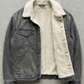 A washed black denim jacket with  a off-white shearling inner lining, laid flat on a textured concrete surface.