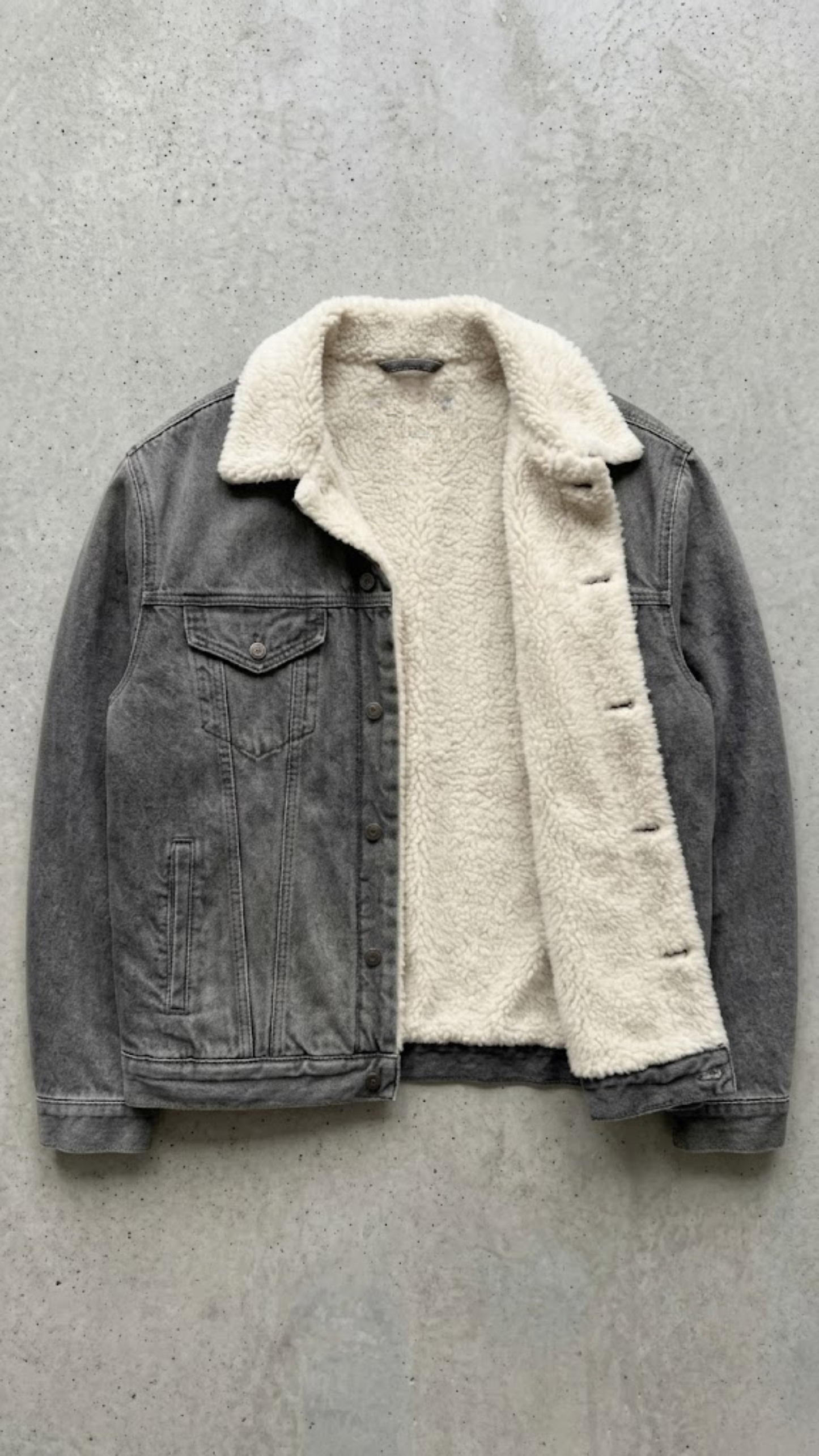 A washed black denim jacket with  a off-white shearling inner lining, laid flat on a textured concrete surface.