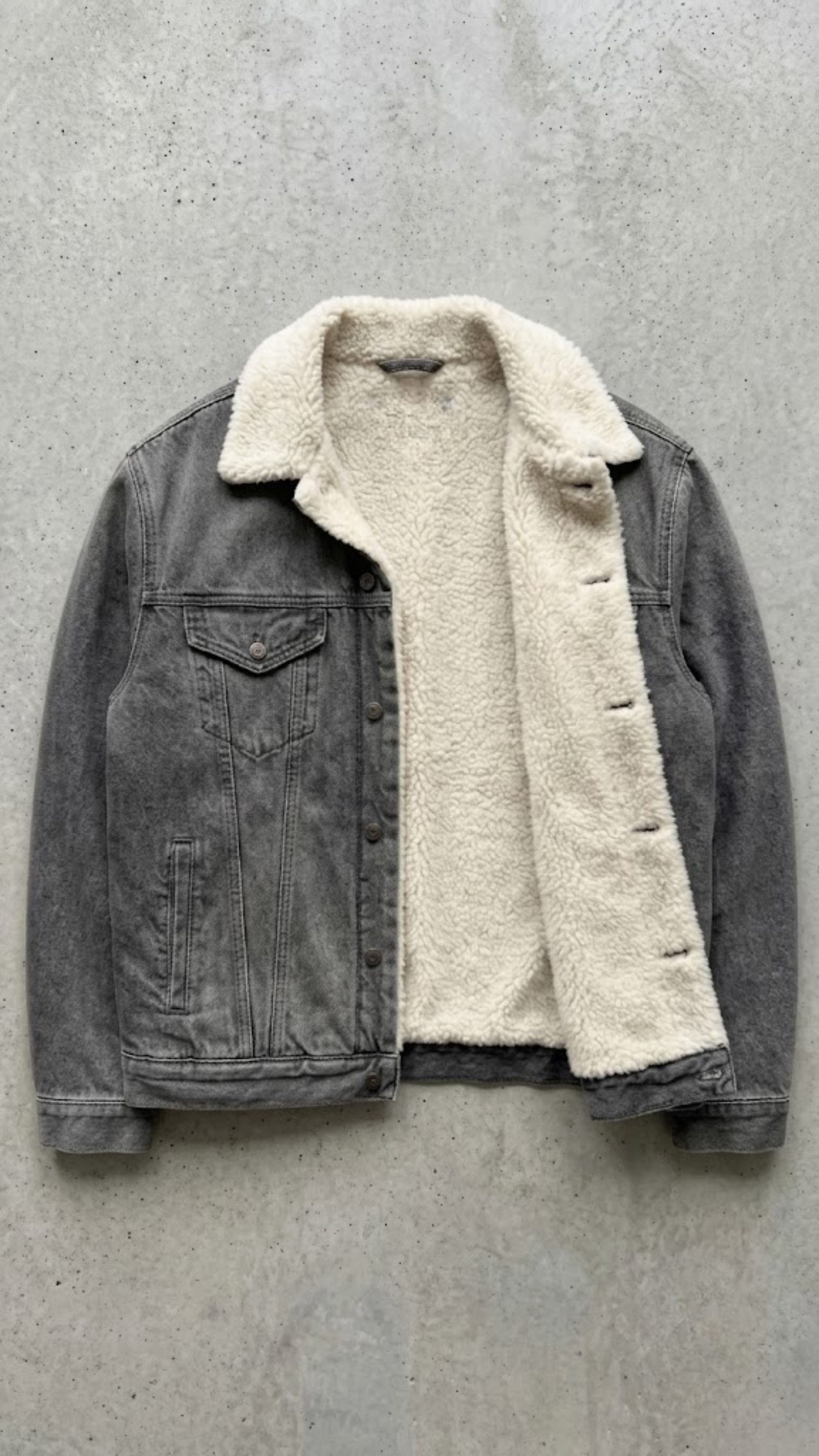 A washed black denim jacket with  a off-white shearling inner lining, laid flat on a textured concrete surface.