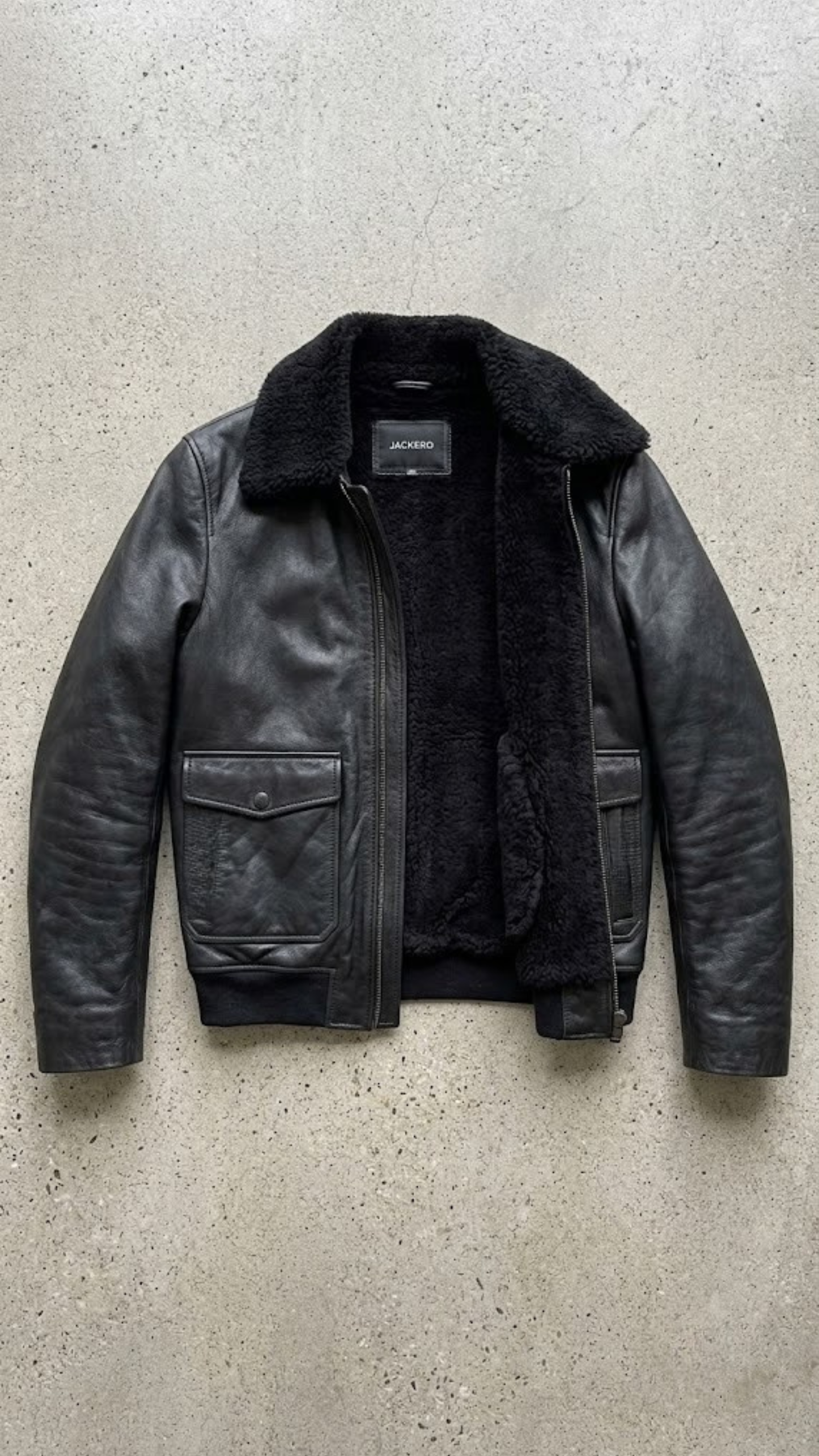 Jackero jacket flat lay photography of a high-end black leather flight jacket with shearling lining on a solid grey backdrop.