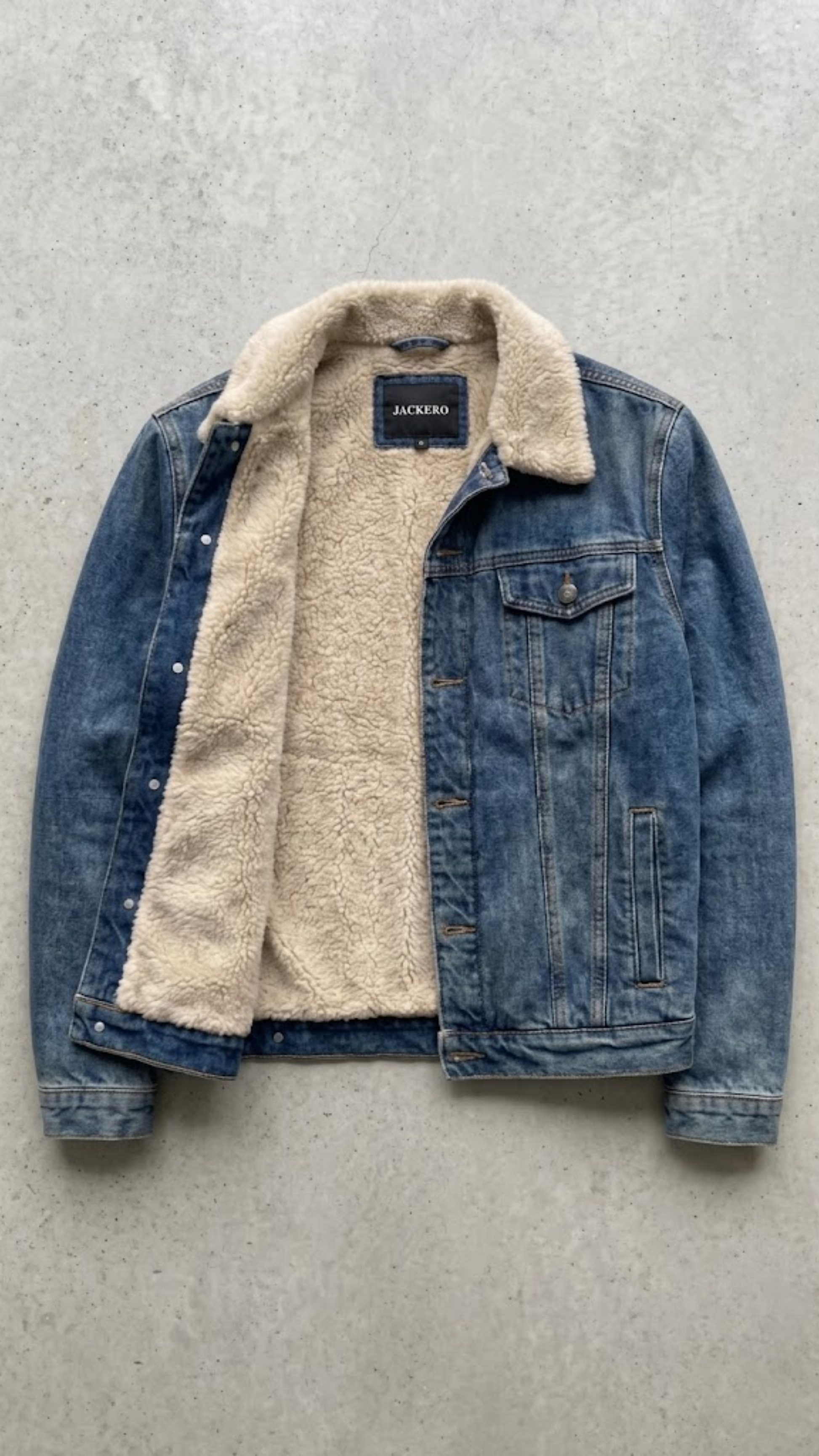A dark gray washed denim jacket with a thick cream-colored sherpa lining, laid flat against a textured gray concrete background.