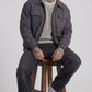 Man sitting on a wooden stool wearing a gray suede jacket, sweater, and jeans against a white background