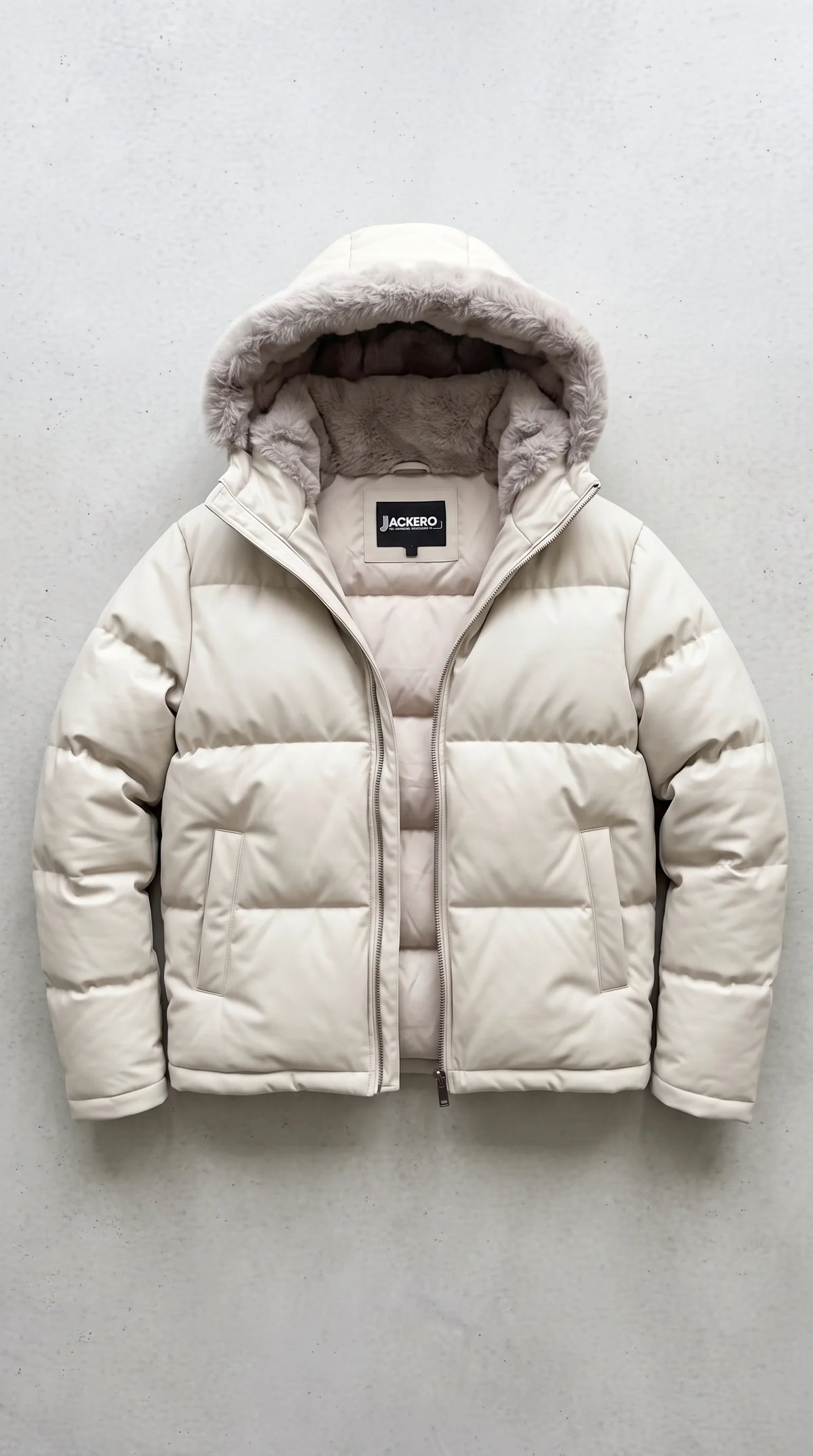A cream-colored puffer jacket with a plush faux-fur trimmed hood, laid flat against a textured gray background. The brand label 'JACKERO' is visible on the inner neck tag.