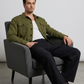 Man sitting on a gray chair wearing a green cotton jacket and dark pants against a plain background