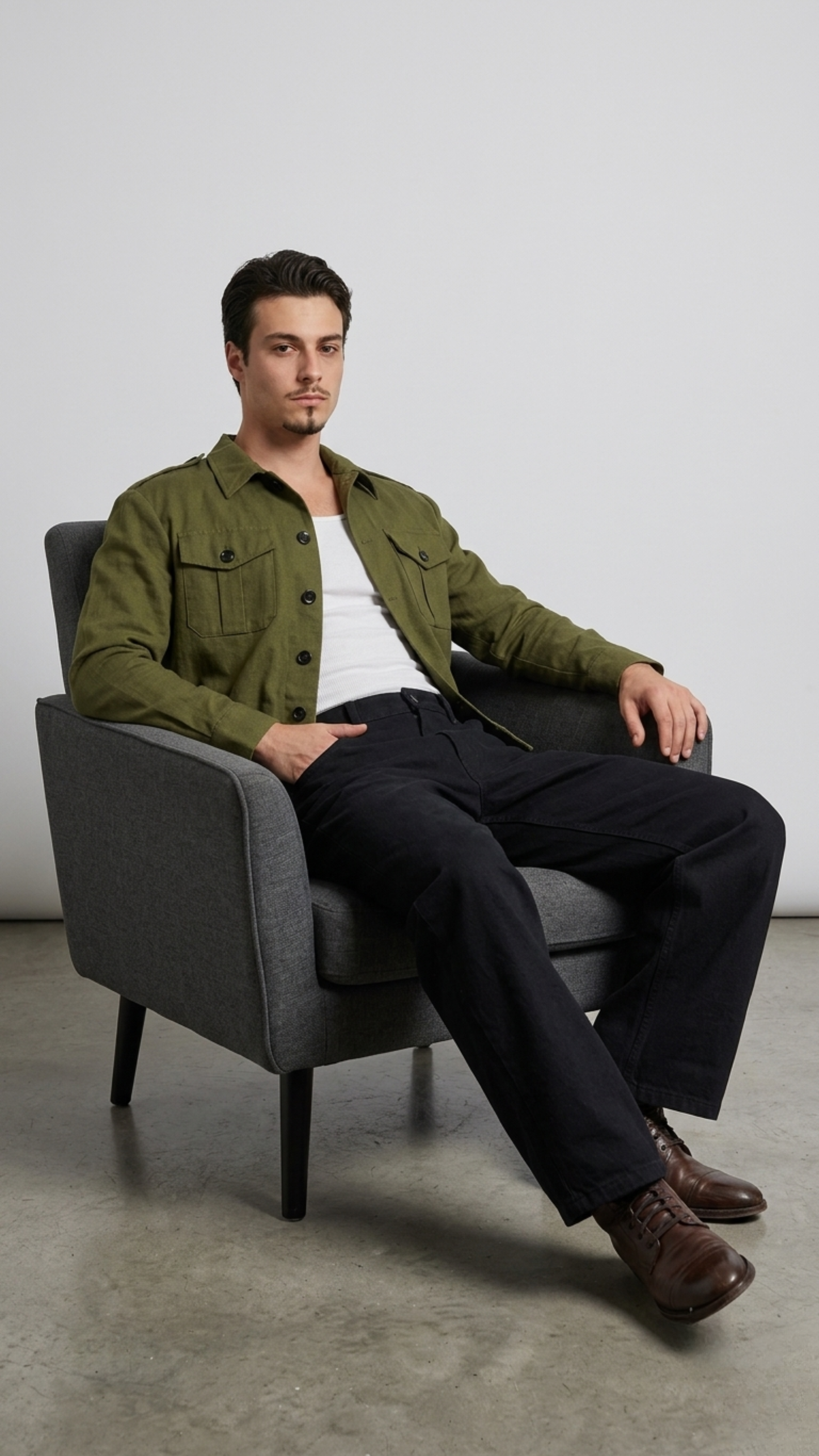 Man sitting on a gray chair wearing a green cotton jacket and dark pants against a plain background