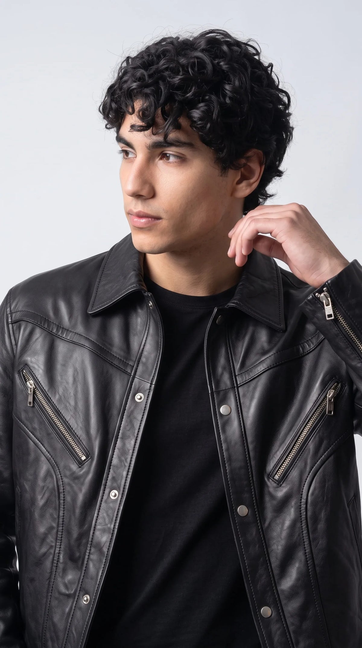 Close-up of a jet-black genuine leather jacket featuring two angled chest pockets with polished metal zippers, a classic pointed collar and a row of silver snap buttons along the front placket. The softly reflective leather grain and sweeping curved seams give this oversized men's jacket a distinctive architectural look, combining practical pocket design with refined biker-inspired detailing.