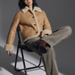 Person wearing a light beige leather coat with plush cream shearling sitting on a chair against a gray background.
