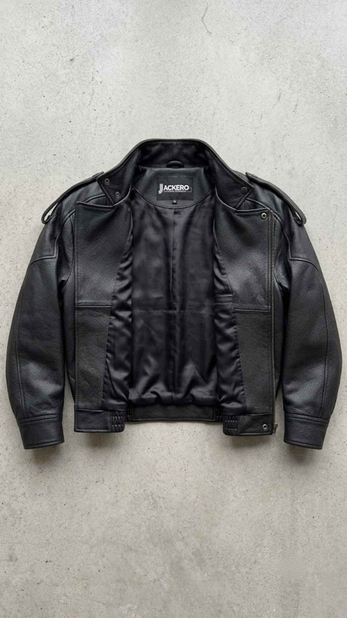 A black leather jacket with a zippered front and ribbed collar, laid flat on a textured gray concrete surface. The brand name 'JACKERO' is visible on the inner neck label.
