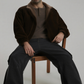 Man sitting on a chair wearing a dark brown suede jacket with fur collar and dark pants against a plain background