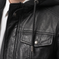 Close-up of a person wearing a black leather jacket with a hood, featuring a drawstring hood, front zipper, chest flap pocket with a snap closure, and a white shirt underneath.