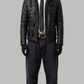 A man modeling a black leather jacket over a white shirt, black tie, and dark jeans against a solid gray background.