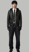 A man modeling a black leather jacket over a white shirt, black tie, and dark jeans against a solid gray background.