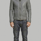 A man wearing a gray leather jacket, dark gray jeans, and brown boots, standing against a solid gray background.