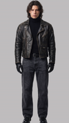 Man wearing a black leather jacket, black turtleneck, jeans, and gloves on a gray background