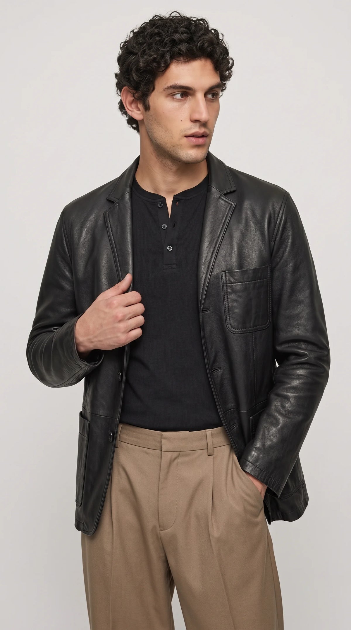 A men's genuine black leather blazer with classic notch lapels, a structured tailored profile and a practical chest pocket is styled with a black Henley shirt and beige trousers. The contrast between dark leather and neutral tailoring creates a sophisticated smart-casual outfit for modern city wear, dinner occasions and understated luxury menswear.