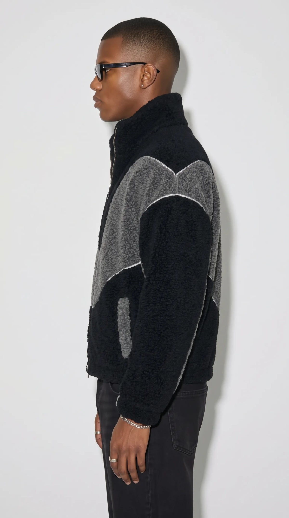 Men’s baggy black and dark grey zip-up fleece jacket with a raised collar, plush textured surface and contrasting grey shoulder panels outlined with white piping. Its relaxed cut and bold graphic construction create a contemporary Y2K outfit with a sporty edge, ideal for pairing with washed black denim, casual trousers and understated everyday essentials.