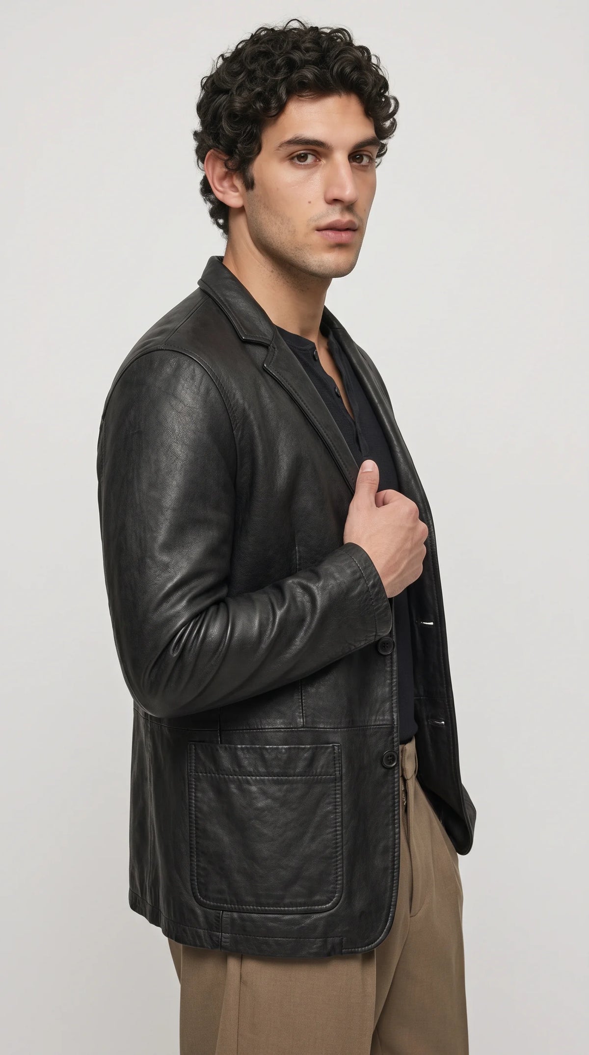 A tailored black leather blazer combines a streamlined shoulder line, classic lapel collar, button-front fastening and neatly finished side welt pockets. Styled with a dark Henley top and warm beige trousers, this versatile leather jacket blends refined tailoring with relaxed urban fashion for creative workplaces, evening outings and premium everyday streetwear.
